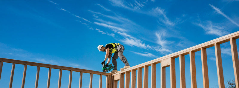 A construction worker in a high-visibility yellow vest and white hard hat is installing or adjusting a green power tool on a wooden railing outdoors under a bright blue sky with wispy clouds.